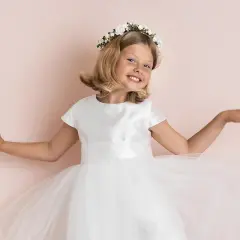 A smiling young girl in a white dress and flower crown, with arms outstretched.