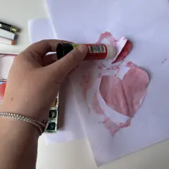 Mano che incolla un cuore di carta su un foglio, creando bellissimi disegni per la mamma. Vicino, acquerelli e pennarelli.