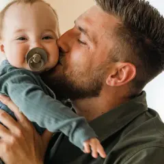 Un padre bacia teneramente il suo bambino appena nato, che sorride con un ciuccio.