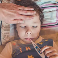 Bambino con febbre, mano sulla fronte, termometro in bocca. Possibili sintomi streptococco bambini.