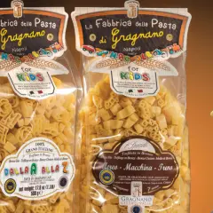 Due confezioni di pasta per bambini, una con lettere e l'altra con aerei, macchine e treni, prodotte a Gragnano.