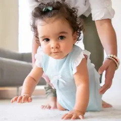 A baby in a blue striped outfit crawls on a white rug, with a person's hands gently supporting them.