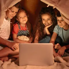 Quattro bambini guardano un film per ragazzi di 12 anni su un laptop, accoccolati in una tenda con luci scintillanti e popcorn.