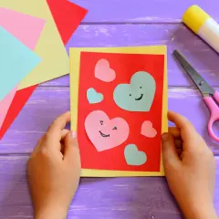 Bambino crea un biglietto con cuori colorati, forbici rosa e colla. Perfetto per lavoretti per bambini creativi.