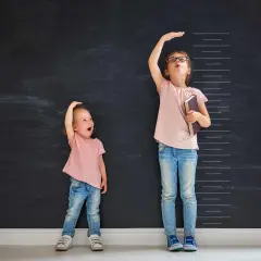 Due bambine misurano la loro altezza su una lavagna. Una, con un'espressione sorpresa, sembra avere l'altezza di un bambino di 2 anni.