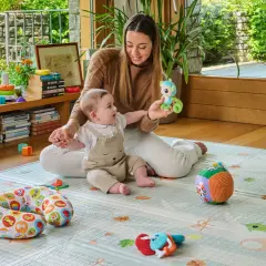 Mamma gioca con neonato 6 mesi che non sta seduto da solo. Il bimbo è su un tappeto gioco con tanti giocattoli.