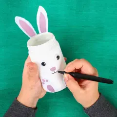 Mani che creano lavoretti di Pasqua bellissimi: un barattolo trasformato in coniglietto con orecchie rosa e zampe glitterate.