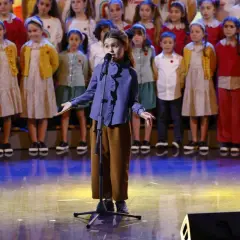 Una bambina canta sul palco, circondata da un coro di bambini, un momento che ricorda le 25 canzoni più famose dello Zecchino d'Oro.