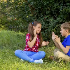 Bambini seduti sull'erba, intenti in divertenti giochi con le mani, ridono felici sotto gli alberi.