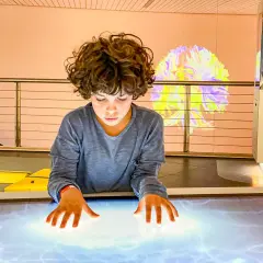 Bambino esplora un tavolo interattivo in uno dei musei per bambini, con un'installazione luminosa a forma di cervello sullo sfondo.