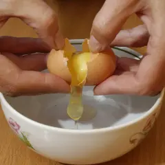 Mani che rompono un uovo per preparare uova alla coque, con il tuorlo che cola in una ciotola.