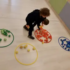 Bambini 3-5 anni impegnati in giochi motori, spostano palline colorate tra cerchi sul pavimento.