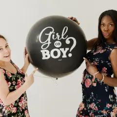 Due donne sorridono tenendo un palloncino nero con scritto "Girl or Boy?". Il **gender reveal significato** è l'attesa di una nuova vita.