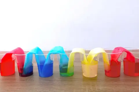 Colorati esperimenti per bambini: strisce di carta assorbono liquidi colorati in bicchieri trasparenti, creando un arcobaleno liquido.