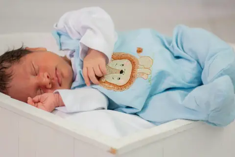 A newborn baby sleeps peacefully in a white bassinet, wearing a light blue onesie with a lion embroidery.