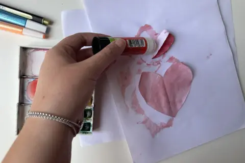Mano che incolla un cuore di carta su un foglio, creando bellissimi disegni per la mamma. Vicino, acquerelli e pennarelli.