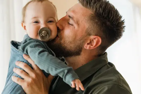 Un padre bacia teneramente il suo bambino appena nato, che sorride con un ciuccio.