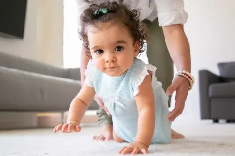 A baby in a blue striped outfit crawls on a white rug, with a person's hands gently supporting them.