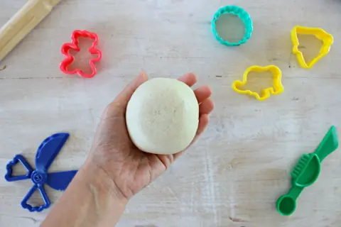 Mano che tiene una palla di pasta di sale, pronta per creare forme con stampini colorati e attrezzi. Ricetta facile per divertirsi.