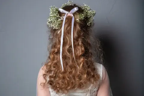 Capelli ricci con coroncina di fiori e nastri, perfetti per trecce acconciature comunione.