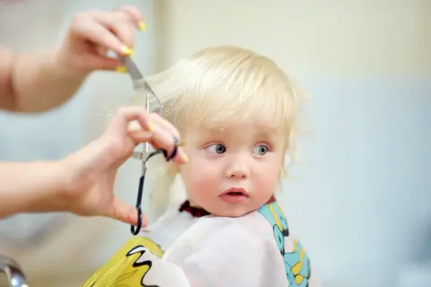 Bambino biondo con occhi azzurri riceve il suo primo taglio capelli, un momento speciale immortalato con cura.