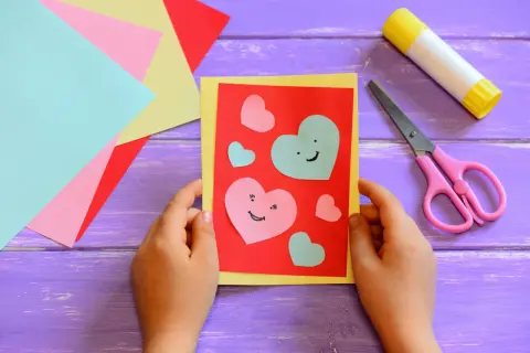 Bambino crea un biglietto con cuori colorati, forbici rosa e colla. Perfetto per lavoretti per bambini creativi.