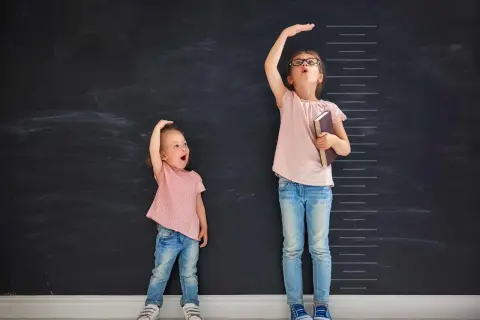 Due bambine misurano la loro altezza su una lavagna. Una, con un'espressione sorpresa, sembra avere l'altezza di un bambino di 2 anni.