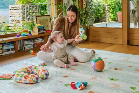 Mamma gioca con neonato 6 mesi che non sta seduto da solo. Il bimbo è su un tappeto gioco con tanti giocattoli.