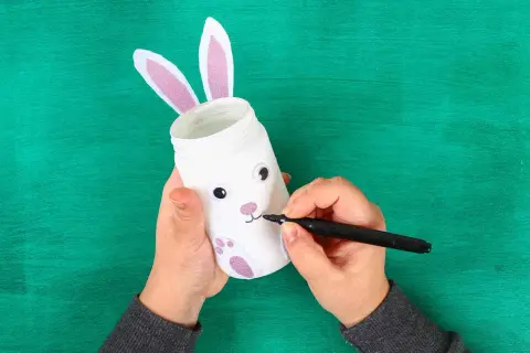 Mani che creano lavoretti di Pasqua bellissimi: un barattolo trasformato in coniglietto con orecchie rosa e zampe glitterate.