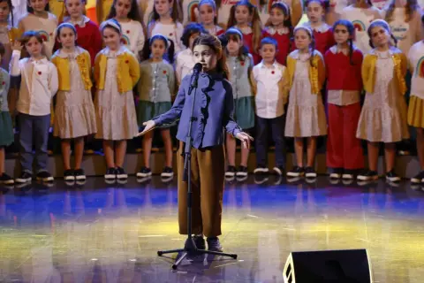 Una bambina canta sul palco, circondata da un coro di bambini, un momento che ricorda le 25 canzoni più famose dello Zecchino d'Oro.