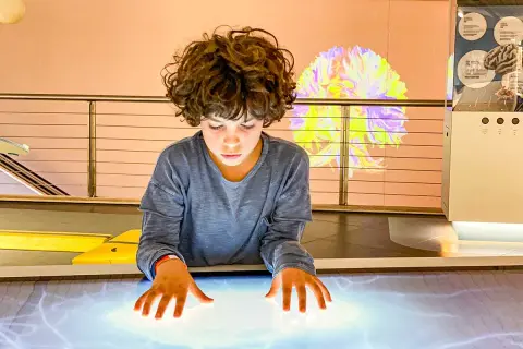 Bambino esplora un tavolo interattivo in uno dei musei per bambini, con un'installazione luminosa a forma di cervello sullo sfondo.