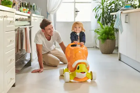 Un papà sorridente incoraggia i primi passi del suo bambino su un cavalcabile a forma di tartaruga in cucina.