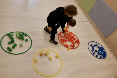 Bambini 3-5 anni impegnati in giochi motori, spostano palline colorate tra cerchi sul pavimento.