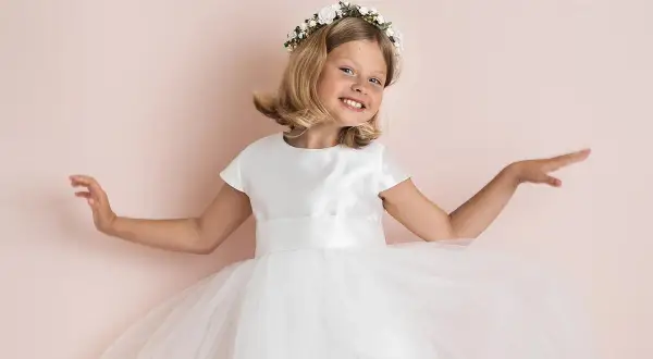 A smiling young girl in a white dress and flower crown, with arms outstretched.