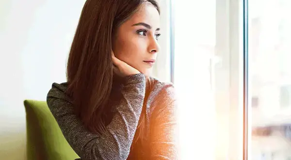 Donna pensierosa guarda fuori dalla finestra, preoccupata per un ciclo in ritardo e come farlo venire.