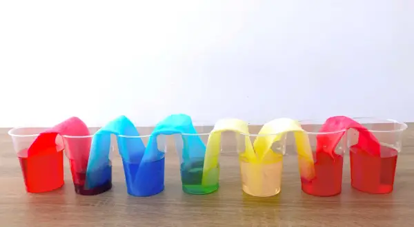 Colorati esperimenti per bambini: strisce di carta assorbono liquidi colorati in bicchieri trasparenti, creando un arcobaleno liquido.