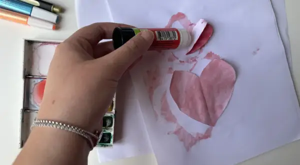 Mano che incolla un cuore di carta su un foglio, creando bellissimi disegni per la mamma. Vicino, acquerelli e pennarelli.