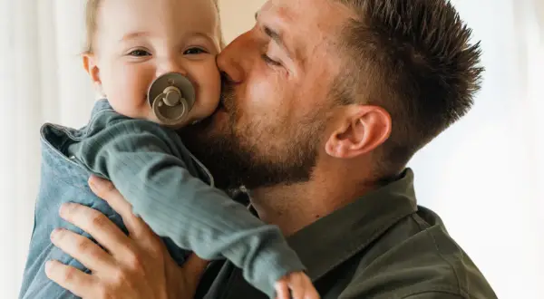 Un padre bacia teneramente il suo bambino appena nato, che sorride con un ciuccio.