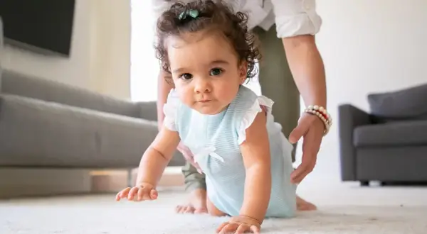 A baby in a blue striped outfit crawls on a white rug, with a person's hands gently supporting them.