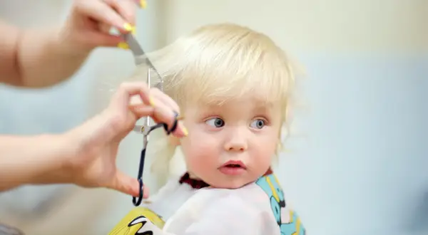 Bambino biondo con occhi azzurri riceve il suo primo taglio capelli, un momento speciale immortalato con cura.