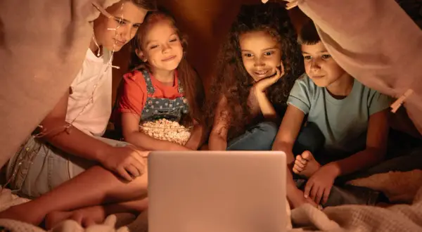 Quattro bambini guardano un film per ragazzi di 12 anni su un laptop, accoccolati in una tenda con luci scintillanti e popcorn.