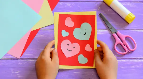 Bambino crea un biglietto con cuori colorati, forbici rosa e colla. Perfetto per lavoretti per bambini creativi.
