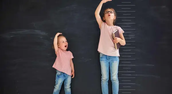 Due bambine misurano la loro altezza su una lavagna. Una, con un'espressione sorpresa, sembra avere l'altezza di un bambino di 2 anni.
