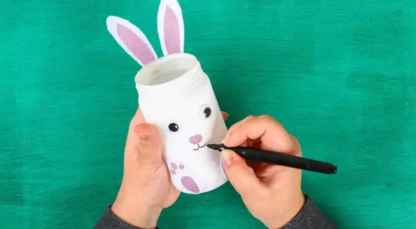 Mani che creano lavoretti di Pasqua bellissimi: un barattolo trasformato in coniglietto con orecchie rosa e zampe glitterate.