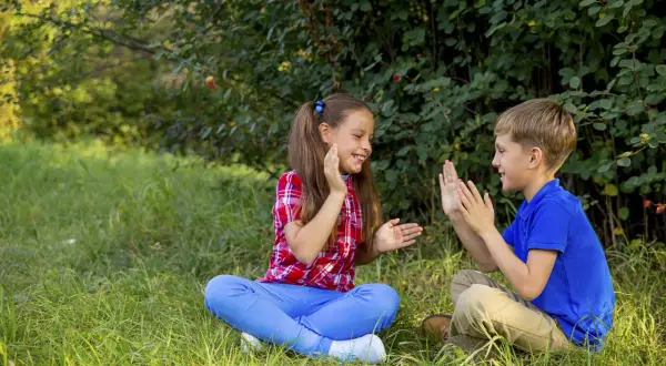 Bambini seduti sull'erba, intenti in divertenti giochi con le mani, ridono felici sotto gli alberi.