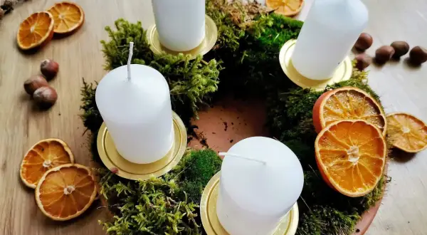 Corona dell'avvento fai da te con muschio, candele bianche, fette d'arancia essiccata e nocciole.