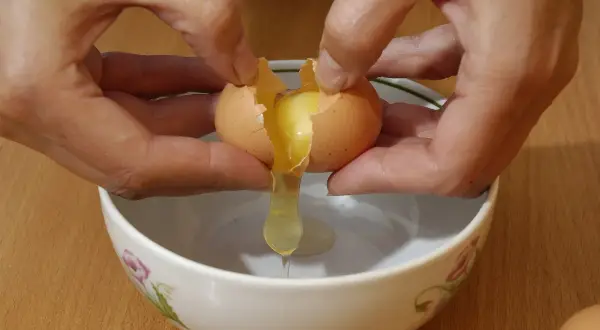 Mani che rompono un uovo per preparare uova alla coque, con il tuorlo che cola in una ciotola.