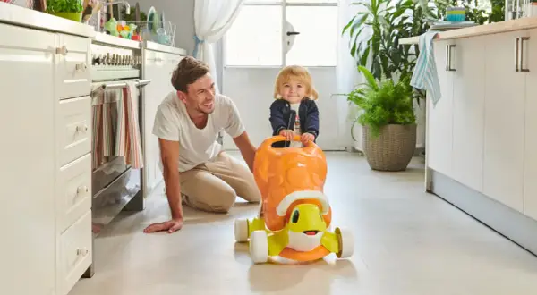 Un papà sorridente incoraggia i primi passi del suo bambino su un cavalcabile a forma di tartaruga in cucina.