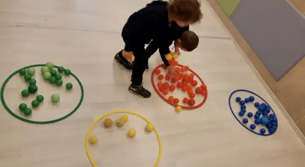 Bambini 3-5 anni impegnati in giochi motori, spostano palline colorate tra cerchi sul pavimento.