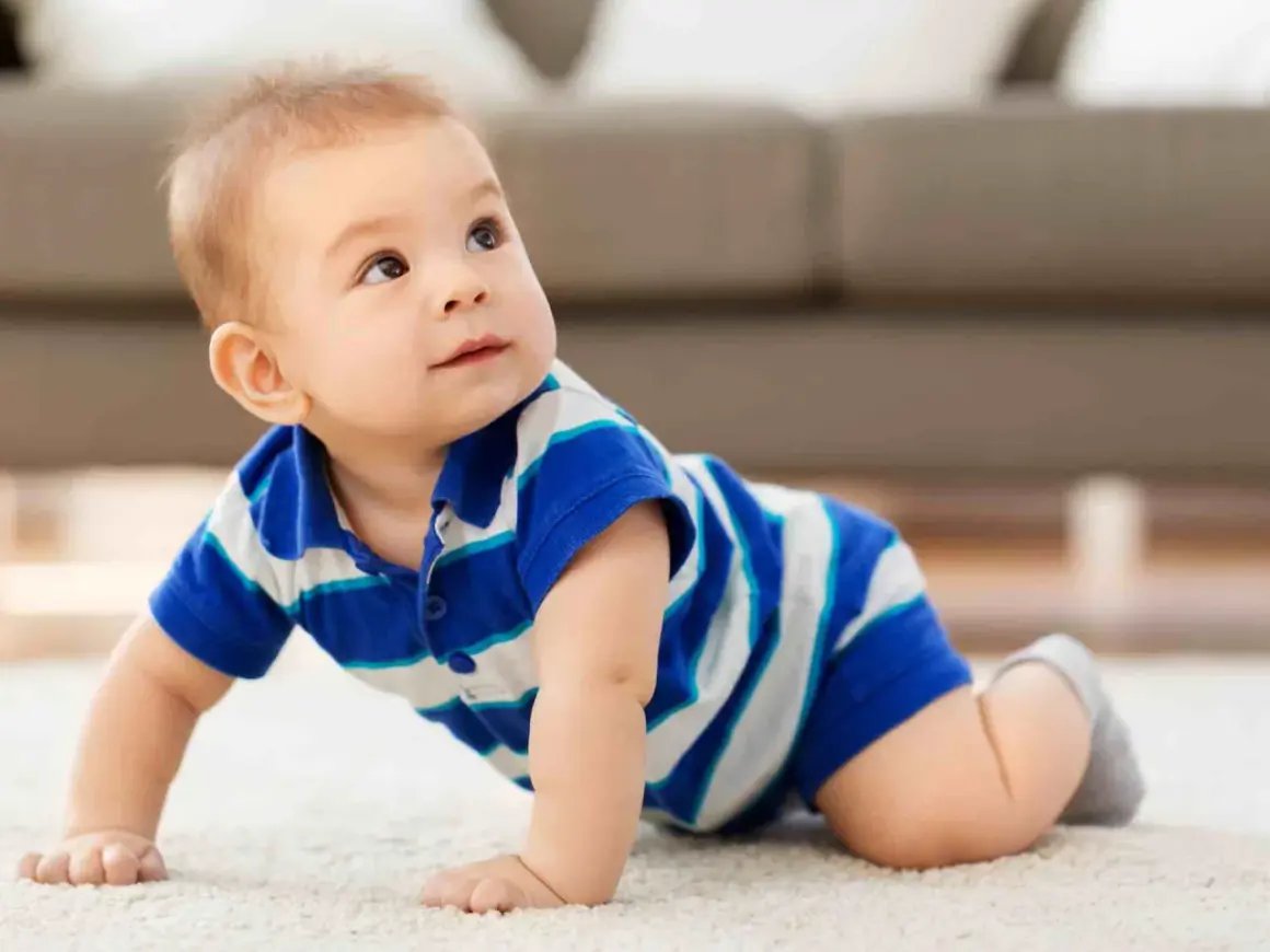 Un adorabile neonato di 8 mesi, con indosso una maglietta a righe blu e bianche, gattona curioso su un tappeto chiaro, esplorando il mondo.